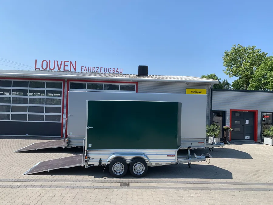 Zwei Anhänger verschiedener Größen stehen nebeneinander vor der LOUVEN Fahrzeugbau-Werkstatt. Im Hintergrund ist die Werkstatt mit roten Rahmen und Glasfront zu sehen.